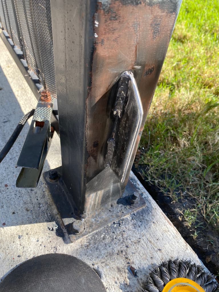 Fresh weld on a steel gusset reinforcing the base of a gate post, with a grinder disc and wire wheel nearby